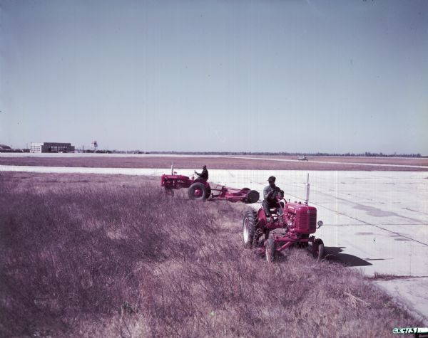 Slightly elevated view of two men using McCormick Farmall Super A tractors to mow grass near the runway of an airport. The original caption reads: "This is the main airline field serving Savannah.  It has 2800 acres of which 1500 must be mowed to keep the field in first class shape.  Previous mowing was by low speed units and required 30 days to completely cover the field, resulting in many sections skipped due to lack of time.  These new mowers will travel 22 miles per hour compared to the 7 mph for the older models.  Frank Lehardy says he will get over the entire field with this new equipment in a few days.  Never spending more than 1/4 of the time they formerly needed. Statement by airport manager, Frank Lehardy, 'Lower costs, less manpower and a better kept field are the results we expect from this new fast equipment.'"