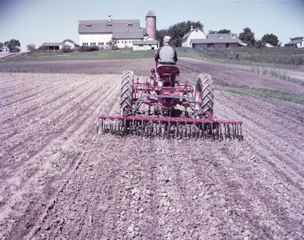 View towards a man using a McCormick Farmall Super C with a C-254 cultivator in a field. Barns and farm buildings are in the background. The original caption reads: "C-254 two-row cultivator with No.5 (10-foot) weeder-mulcher and Farmall Super C tractor. First cultivation in corn on IH Hinsdale farm."