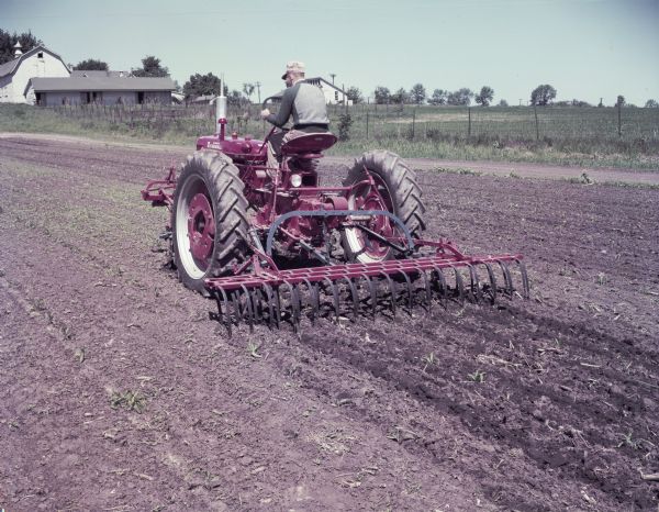 View towards a man using a McCormick Farmall Super C with a C-254 cultivator to work in a field. Barns and farm buildings are in the background. The original caption reads: "C-254 two-row cultivator with No.5 (10-foot) weeder-mulcher and Farmall Super C tractor. First cultivation of corn on IH Hinsdale farm."