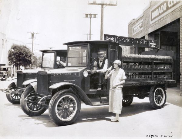 A woman wearing a dress and a cloche hat is standing next to an Orange Crush truck while drinking from a soda bottle. The driver of the truck is sitting behind the wheel looking down at her. He is wearing a straw hat, bow tie, vest, and eyeglasses, Another truck and driver are in the background, and a building with a sign for the Dorado Club is on the right. The text on the truck reads: "Drink Orange Crush in Krinkly Bottles; Dorado Club Beverage Co."