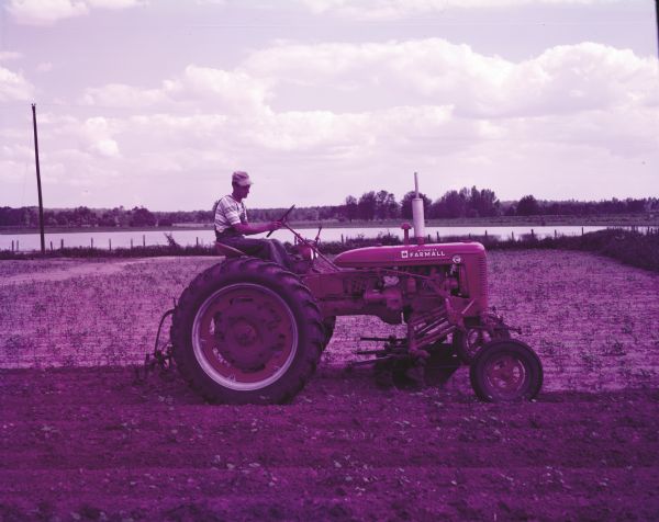 View across field towards J.A. Coon with a McCormick Farmall Super C tractor and a C-254 cultivator. There is a pond or lake in the background. The original caption reads: "C-254 two-row cultivator and Super C with wide front axle.  First cultivation in cotton on 200 acre farm of J.A. Coon, Star Route, Atmore, Ala."