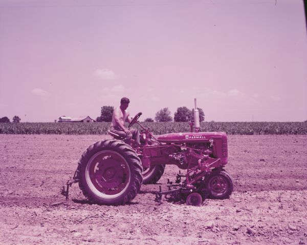 View across field towards a bare-chested man, possibly Raymond Teal, using a Farmall Super C tractor outfitted with a C-254 cultivator to work in a field. A barn is in the background. The original caption reads: "C-254 two-row cultivator with No.10 tooling equipment, straight disk hillers, spring-trip attachment on Super C. First cultivation in cotton on 70 acre farm of Raymond Teal. Rt.4, Albertville, Ala."