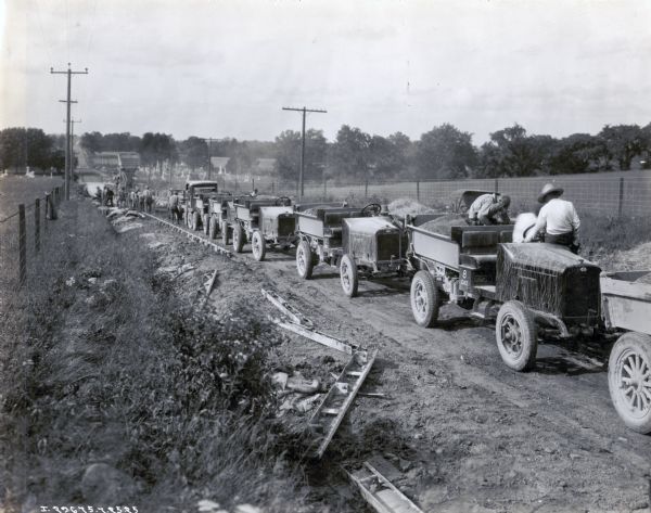 A line of International trucks are parked in a line along a dirt road lined with fences and power lines. Each truck has a load of dirt, and metal forms are staked along the side of the road to hold the new road in place. Men in the background work near what appears to be a cement mixer. There are cemeteries on either the side of the road in the distance.