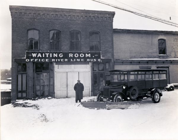 A man wearing a heavy coat, boots, a hat, and eyeglasses, stands outside a brick building marked "Waiting Room. Office River Line Bus Co." An International bus outfitted with a snow plow is parked next to him in the snow.