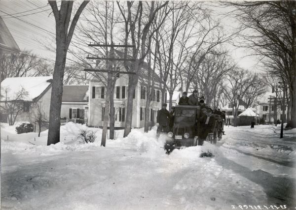 A group of men wearing heavy coats and hats ride on an International truck with snowplow attached. They are driving through heavy snow on a residential street with houses in the background.