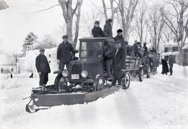 A large group of men wearing winter coats, hats, and gloves stand on or near an International truck outfitted with a snow plow. Men are also sitting and standing in the gated truck bed. The truck is parked on a residential street covered in snow, and there are other trucks behind the snow plow, and houses in the background.