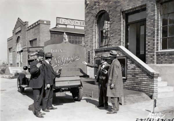 Four men wearing hats are standing next to an International truck while holding bottles of NuGrape soda(?). The lettering on the truck reads: "Drink NuGrape in Bottles, A Flavor You Can't Forget." They are standing in front of a brick building with an obscured sign for NuGrape on the wall of the steps leading up to the entrance.