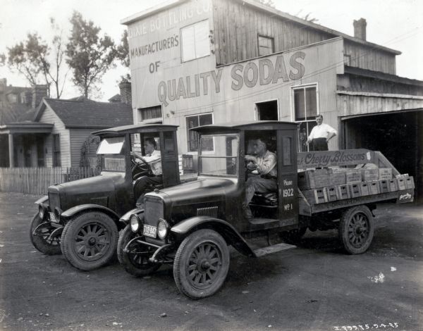 Two men sit in International trucks parked outside the Dixie Bottling Company building. Another man stands in the background, near the building for the bottling company. Houses can be seen on the left. The sign on the back of the foreground truck reads: "Drink Cherry Blossoms, A Blooming Good Drink; Grape-Orange-Lemon Sour and Straw..."