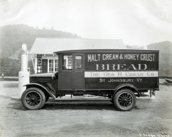 An International truck used by the George H. Cross Company is parked in front of a filling station. In the background is a man standing near railroad tracks. The lettering on the truck reads: "Malt Cream & Honey Crust Bread; The Geo. H. Cross Co., St. Johnsbury, VT."
