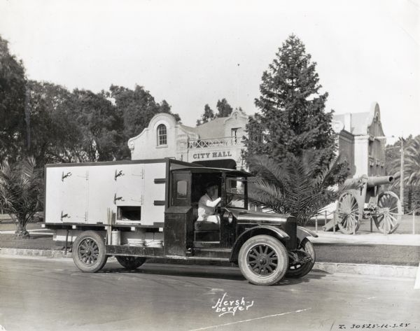 A man sits behind the wheel of an International truck parked in front of an old "City Hall" building. There is a cannon displayed on the front lawn.