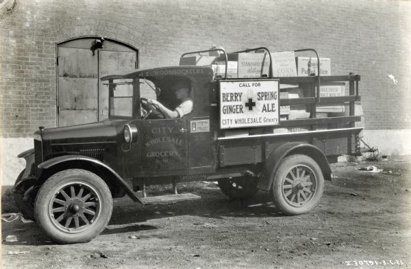 A man is sitting behind the wheel of an International truck used by the City Wholesale Grocery Company. The truck is near the doorway of a brick building, and boxes of cereal are loaded in the back of the truck. The sign on the truck reads: "Call for Berry Ginger Spring Ale, City Wholesale Grocery."