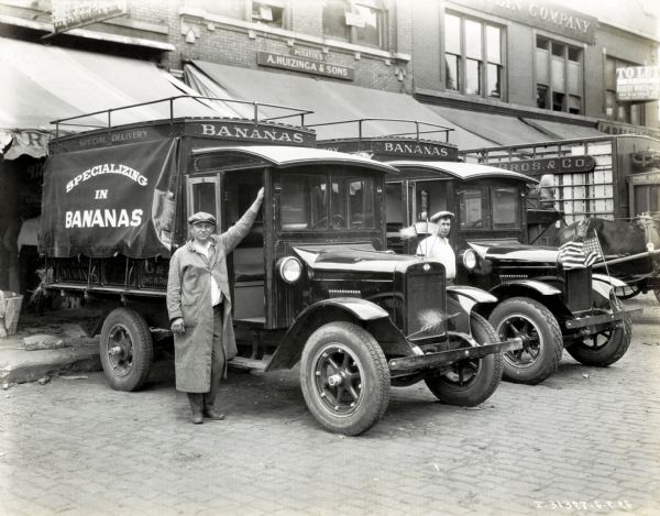 Two men stand next to International trucks marked "Bananas" and decorated with American flags parked outside a commercial building.