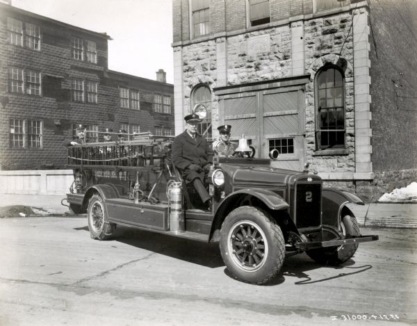 Two men sitting in an International Model SF-34 1927 fire truck marked "Ogdensburg, N.Y." They are parked in front of what appears to be a fire station.