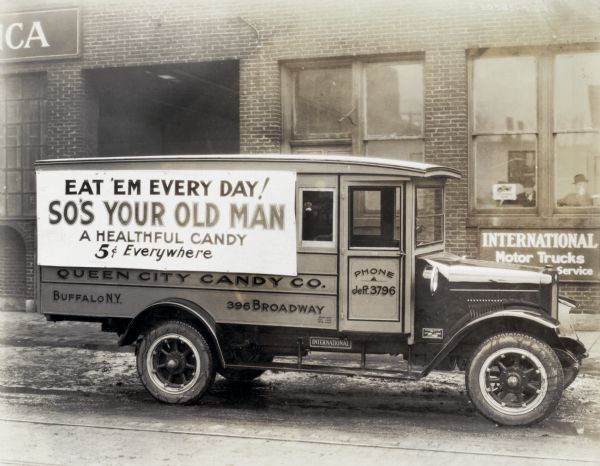 An International truck used by the Queen City Candy Company parked in front of an International motor trucks sales and service office. Two men inside the office are looking out the windows. The text on the truck reads: "Eat 'Em Every Day! So's Your Old Man; A Healthful Candy; 5 Cents Everywhere; Queen City Candy Co.; Buffalo, N.Y. 396 Broadway."