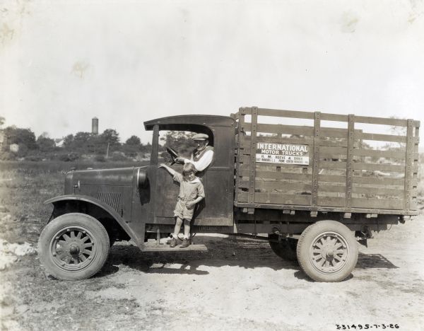 A child stands on the running board of an International truck while a man sits behind the steering wheel. There is a silo or water tower in the background. The sign on the truck reads: "International Motor Trucks; H.M. Reeve & Sons; East Moriches, L.I."