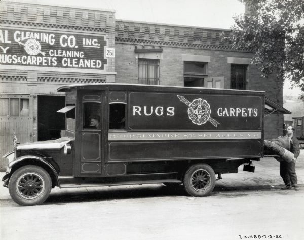 One man is sitting in the cab and another man is standing in the street near the back with a rolled carpet on the bed of an International truck used by the General Cleaning Company. The truck is parked in front of a building with a sign for the General Cleaning Company above an open doorway. The text on the truck reads: "241-251 Maple St. Secaucus, N.J."