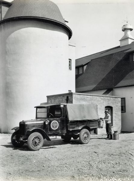 A man loading milk pails into an International truck used by the Yeomen City of Childhood. A large barn with a weathervane on the roof, a silo, and a small brick building are in the background.