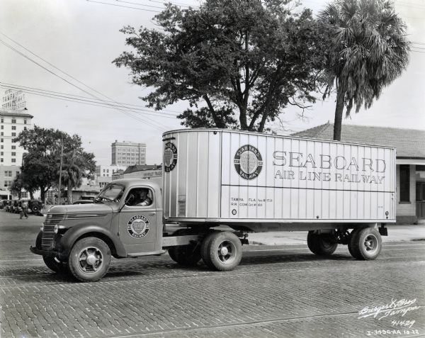 An International truck used by the Seaboard Air Line Railway parked on a brick-paved street. A man is sitting in the cab. Commercial buildings, including the Hotel Tampa Terrace on the left, are in the background.