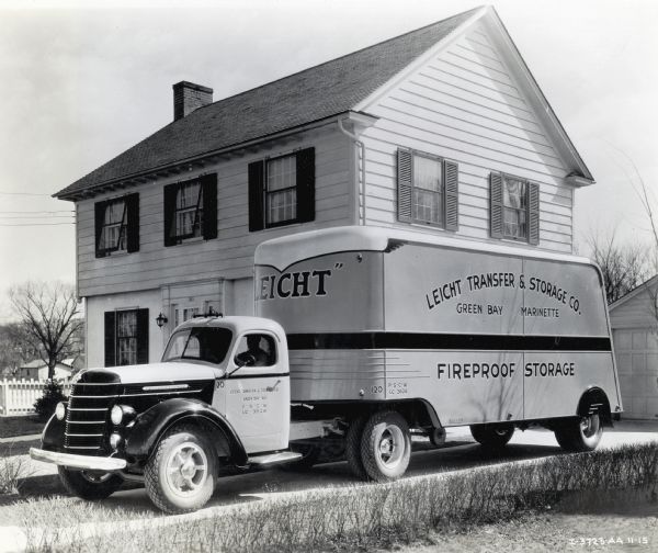 An International truck used by the Leicht Transfer & Storage Co. parked in a driveway beside a residence and a garage. A man is sitting in the cab. The lettering on the truck reads: "Green Bay - Marinette" and "Fireproof Storage."