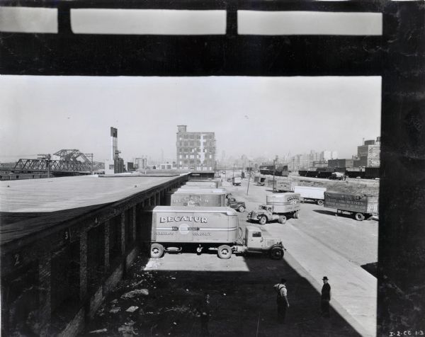 Elevated view through window of multiple International trucks used by the Decatur Cartage Company parked in a loading dock. Two men are standing in the foreground and industrial buildings are in the background. The original caption reads: "Taken from window of Decatur Cartage Company Office showing part of their fleet at their loading dock in Chicago."