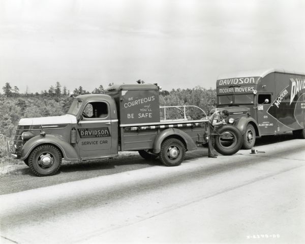 A man changes the tire of a Davidson Transfer & Storage Company truck on the side of a road.  The vehicles shown include an International Model D-15 service truck and a Model D-30 moving van. The text on the truck reads: "Fast Davidson Freight, Service Car" and "Be Courteous and You'll Be Safe." The moving van reads,:"Davidson Modern Movers" and "Agents Allied Van Lines Inc."