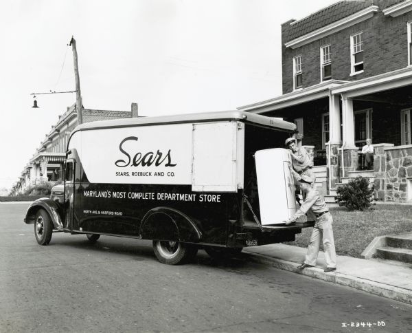 Two uniformed men unload a refrigerator from the back of an International D-30 truck used by the Sears, Roebuck, & Co. truck in front of a residence.  A man wearing a necktie sits on the home's front porch. A line of rowhouses is in the background.
