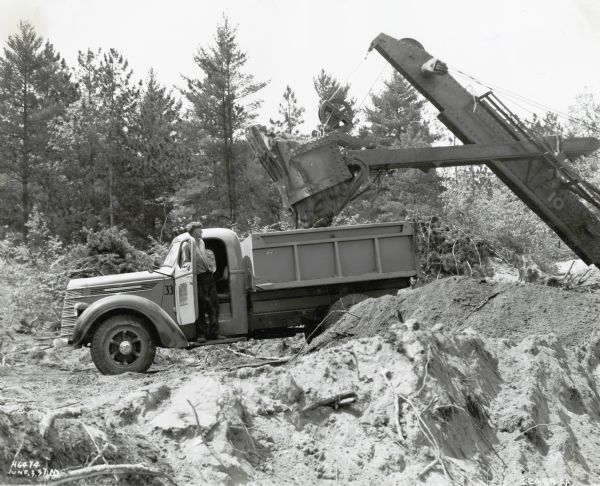 A man loads gravel into the back of an International D-40 truck. The original photograph caption reads: "Removing overburden from Gravel Pit - D40 of 134" w.b. Brennan Paving Co. - Hamilton, Ont.  Also own "C" models." The photograph was taken in Great Bend, Ontario, Canada.