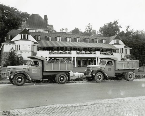 Two International D-40 road repairs trucks used by the Brennan Paving Company are parked on the side of the road with men sitting in the cabs. They are in front of what appears to be a restaurant or resort in Niagara Falls, Ontario, Canada. The original caption reads, "2 D40's, 134" w.b. - Brennan Paving Co., Hamilton, Ontario."