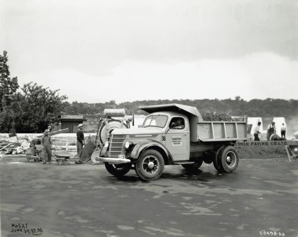 An International D-40 truck used by the Brennan Paving Company is parked in a paved lot in Niagara Falls, Ontario, Canada, while a group of men work in the background. The falls can be seen in the far background. The original caption reads: "Niagara Falls, Ont. Road Repairs; D40 wf. 134" w.b. Brennan Paving Co., Hamilton, Ontario." A "Maid of the Mist" ticket office booth is in the background.
