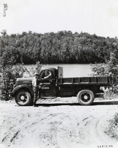 A man in an International D-30 truck used by the Township of Monck is parked in front of a wooded area and body of water. He is talking to a man standing next to the driver's side door.