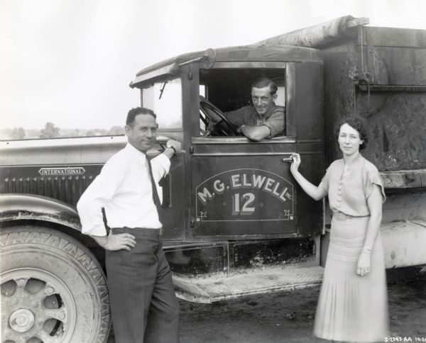 Harold Murphy sits behind the wheel of an International DR-70(?) truck used by contractor M.G. Elwell, left. Mrs. Elwell stands to the right.