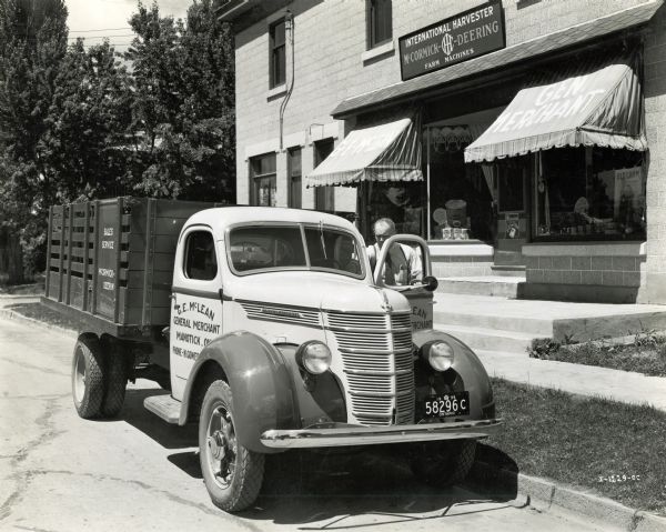 A man is standing at the open door on the driver's side of an International D-30 Special Dealers truck parked outside of an International Harvester agricultural equipment dealership.