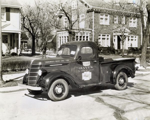 An International D-2 pickup truck used by Hayek Pharmacy parked on a residential street. A man carrying a package is on the front walk of one of the houses behind the truck.