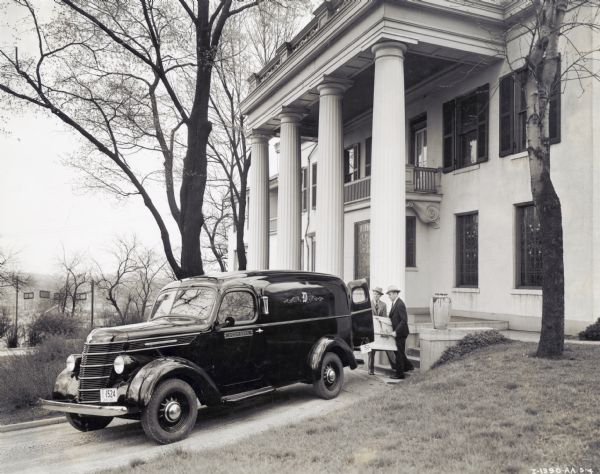 Two men wearing suits and hats are placing a coffin into the back of an International D-2 hearse marked: "Doom Bros." The hearse is parked in front of a large house which has a columned entrance. The house may have served as the Doom Funeral Home.