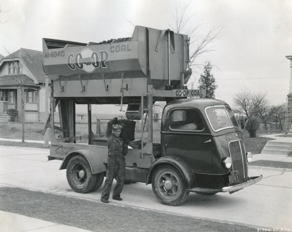 A man hoists a bucket of coal onto his shoulder from a chute in the back of an International D-300 truck owned by Co-op Coal. The truck is parked along a residential street.