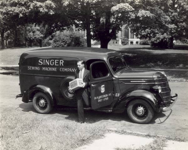 A man carrying a box exits the passenger side of an International D-2 panel truck used by the Singer Sewing Machine Company. The text on the box reads: "Singer H-4 Hand Cleaner."