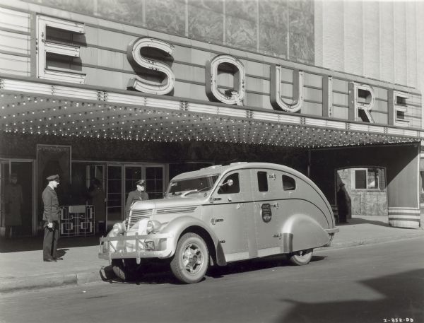 An International D-30 automobile with a special bulletproof body parked outside the Esquire Theater. Two police officers are standing near the vehicle, and a doorman is in the background.