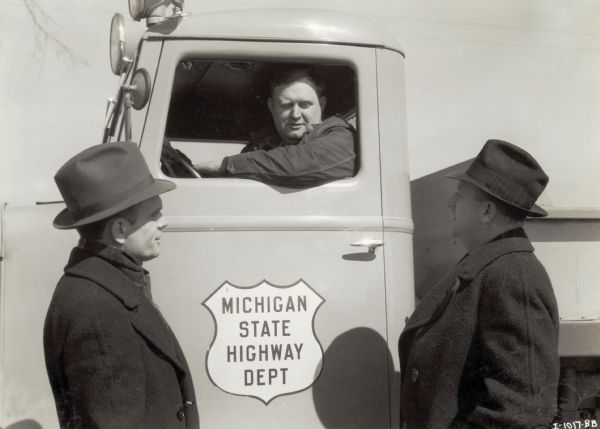 Roman Ederer sits behind the wheel of an International truck marked "Michigan State Highway Department" and speaks to R.C. Richert (left), truck sales manager [dealership or branch house], and H.T. Layster (right), branch manager, who are standing outside by the driver's side door of the truck.