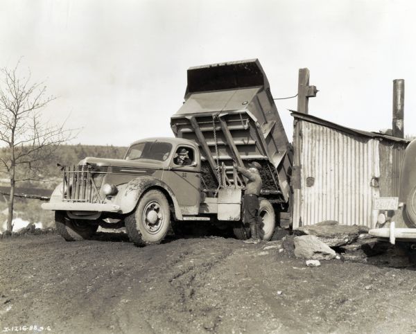 Two men use a DR-60 truck owned by J. Robert Bazley, Inc. to dump coal taken from Pennsylvania coal strippings into a chute leading to railroad cars. The man in the driver's seat is wearing a hat, jacket, and tie. The man standing outside talking to the driver is wearing boots, shirt, trousers, and a hat.