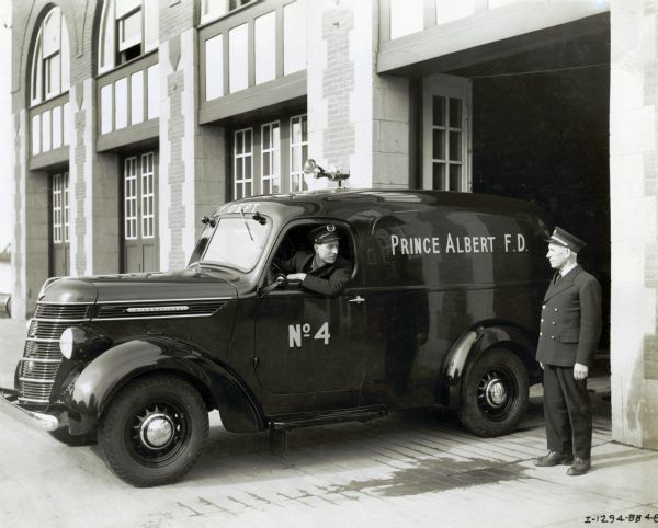 A man is driving an International Model D-2 1938 truck out of the Prince Albert fire department garage while another man is standing nearby. Both men are wearing uniforms with hats.