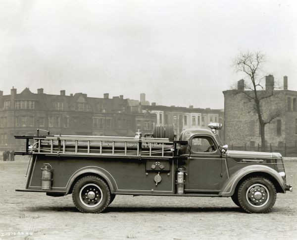 Right side view of an International D-50 Special 750-gallon fire truck with ladders and fire extinguishers mounted on its side parked outdoors. The truck also carries a bell and a fire hose. In the background is a small group of pedestrians, and row houses.
