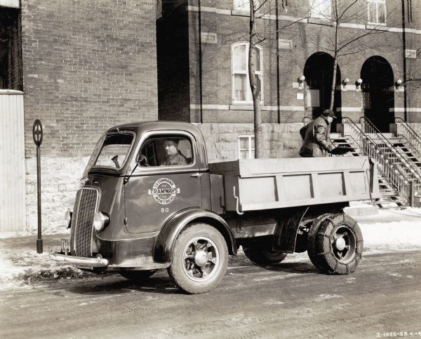 A man is sitting behind the wheel of an International Model D-300 truck used by the Montreal Tramways Company. A man in the back of the truck is holding a shovel. There are brick buildings in the background.
