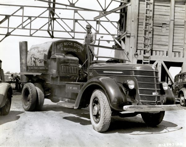 A man stands on the back of an International truck used by the Big Rock Stone & Material Company. The advertisement on the side of the truck features an illustration of an elephant and the text, "Strength! Big Rock Ready-Mixed Concrete." There appears to be a crane or industrial building in the background.