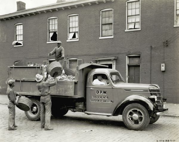 Three men load garbage onto the back of an International DS-35 truck owned by the City of Louisville, while two men sit in the truck's cab. The truck is on a cobblestone road and a brick building with open windows is behind them.