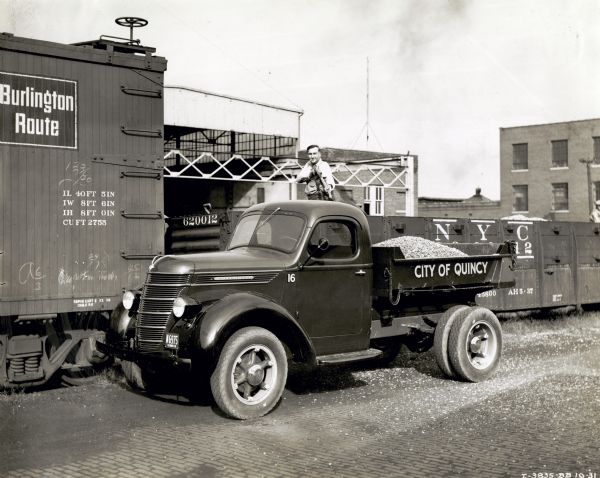 A man smoking a cigarette shovels what appears to be gravel onto the back of an International D-30 truck marked "City of Quincy" from a train car.