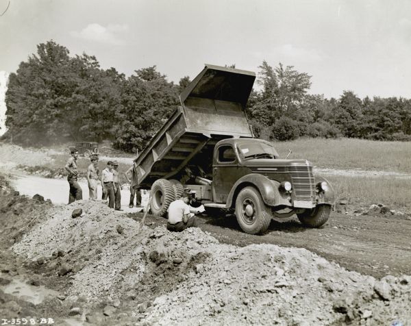 A group of men use an International DR-60 dump truck to work on the New International Bridge Project. There is a car parked in the background.