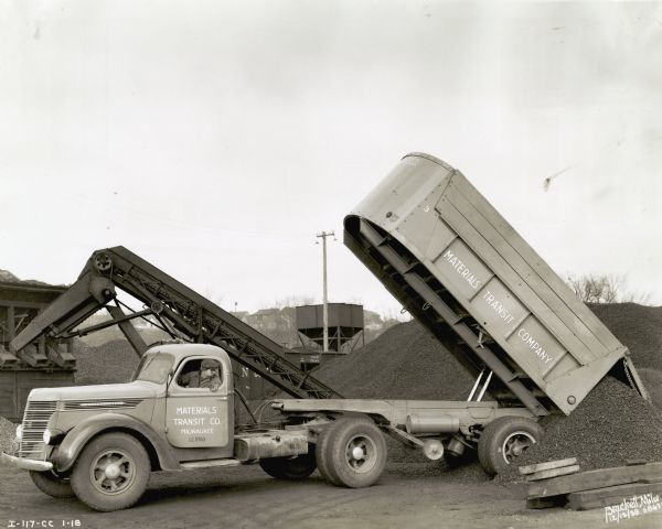 A man from the Material Transit Company drives an International D-50 dump truck to unload what appears to be gravel or rocks. There are houses on a hill in the background.
