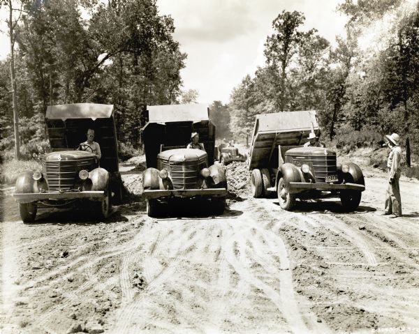 Three men drive International dump trucks down a dirt pathway during the construction of the Natchez Trace Parkway. Another man stands to the side.