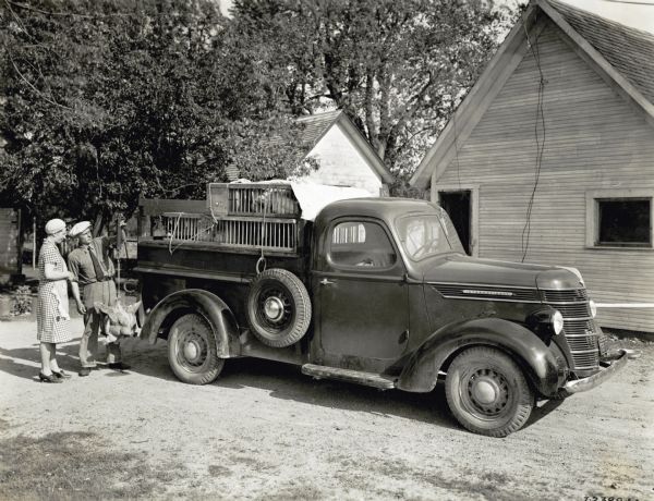 A man and woman stand next to an International pickup truck loaded with caged poultry. There are farm buildings in the background.