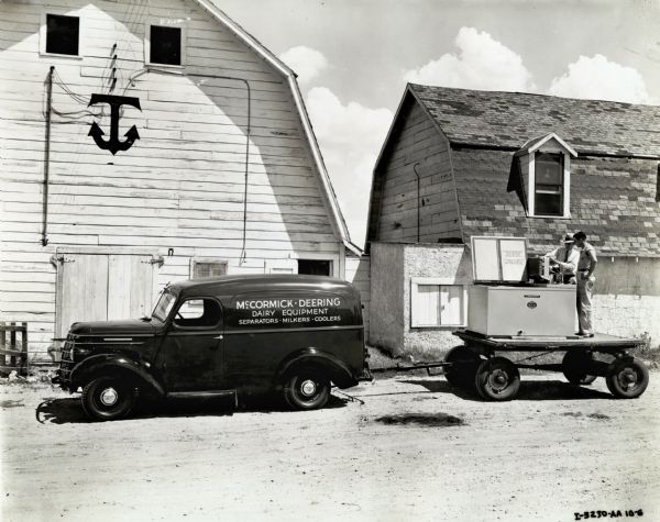 C.L. Thompson, a McCormick-Deering dealer explains a milk cooler, which is loaded on a trailer hauled by an International truck, to Wayne Wakefield. Both men are standing on the back of the trailer. In the background is a small farm building, and a large barn with an anchor symbol on the front.